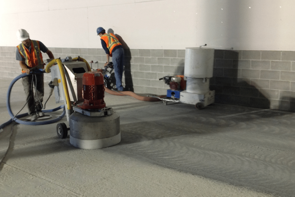 Two construction workers using industrial floor grinders to polish a concrete surface in a commercial space.