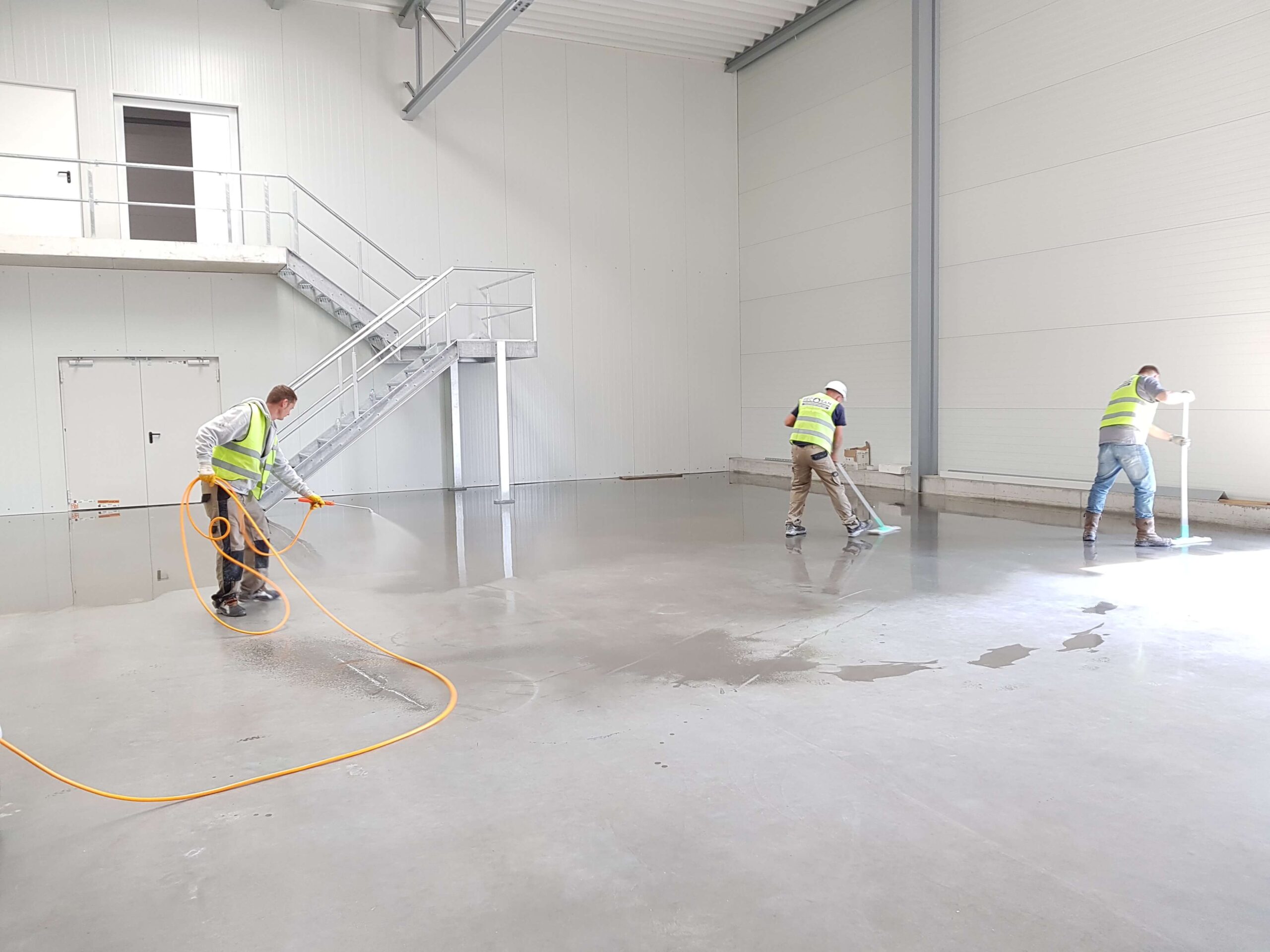 Three technicians in high-visibility vests applying a protective sealant to a large concrete warehouse floor.