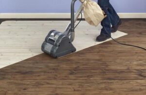 A professional floor sander machine stripping an old dark timber floor to reveal fresh light wood underneath.