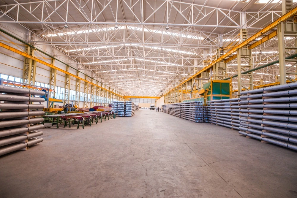 Spacious factory floor with high metal truss ceilings and large stacks of industrial aluminum billets stored on sides.